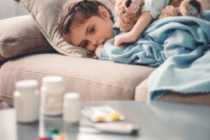 Sick little girl covered in blanket is hugging teddy bear and looking sadly on medicine while lying on couch
